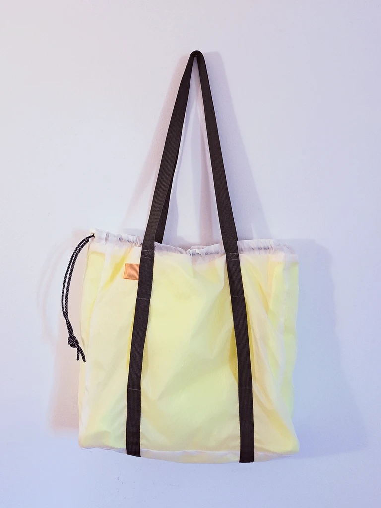 Pale yellow ripstop tote with black straps and a drawstring hanging on a white wall